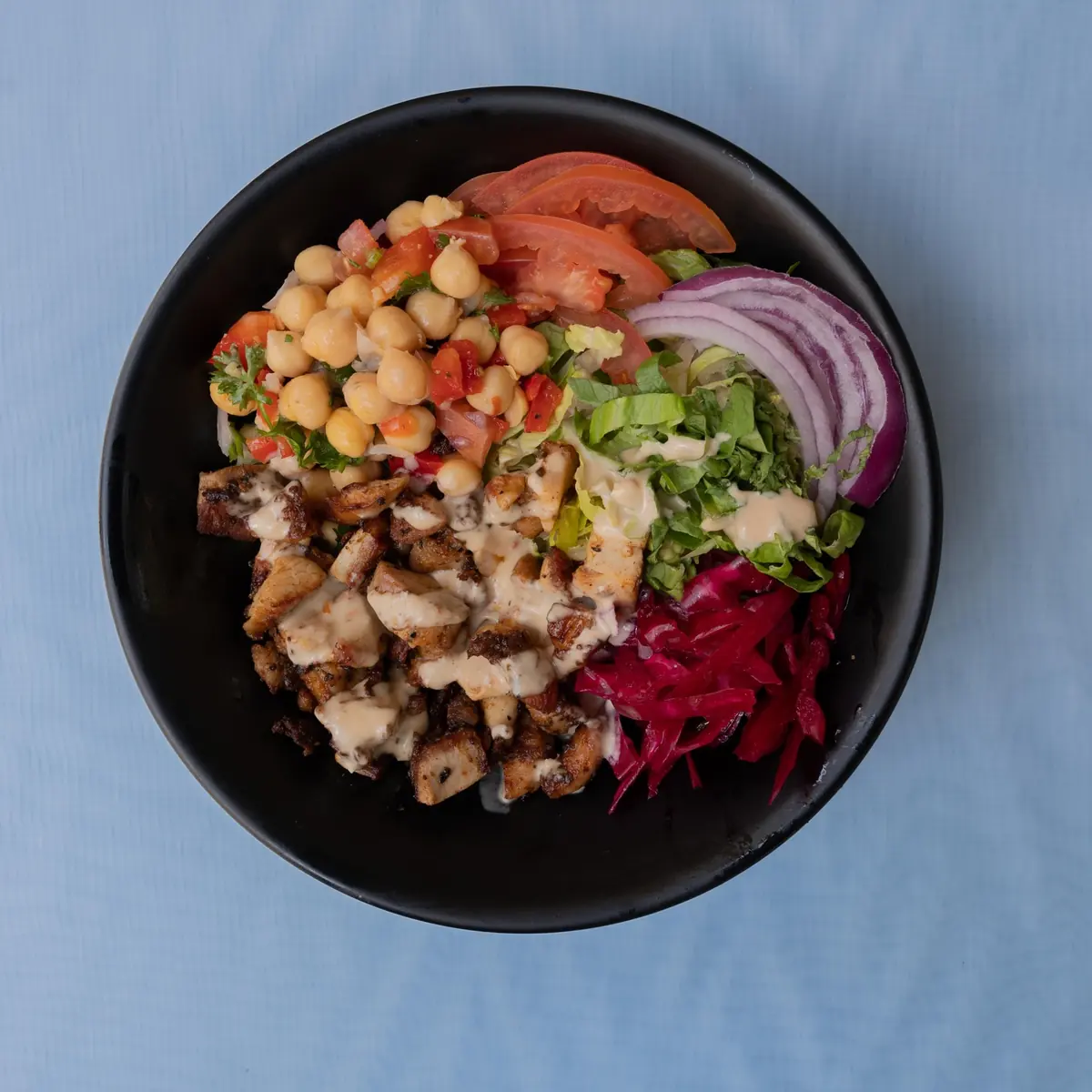 Overhead view of a chicken bowl with chickpeas, fresh vegetables, and sauce at Anatolia Cafe