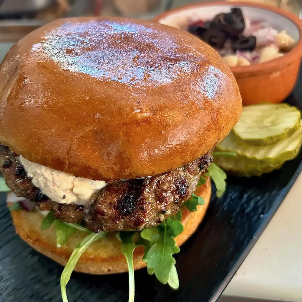 A lamb burger with a glossy bun, lettuce, tomato, and a side of pickles